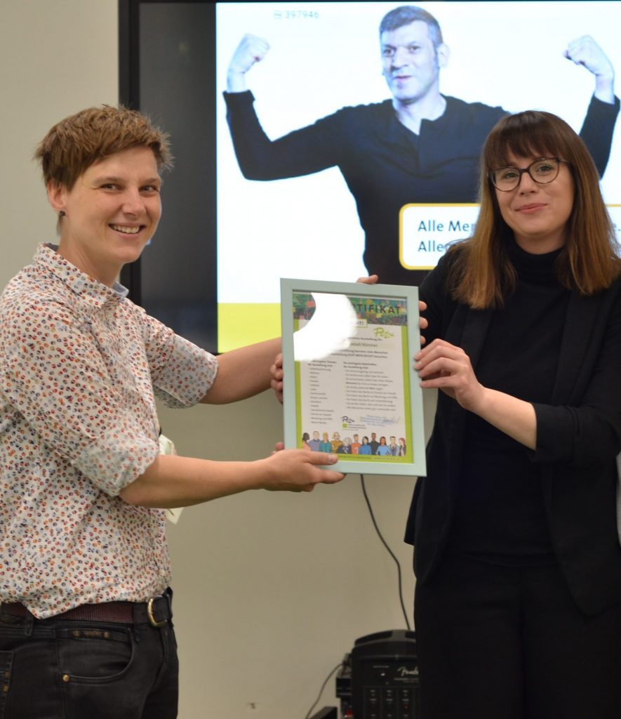 Das Foto zeigt zwei Menschen. Links die Münchner Stadträtin Barbara Likus und rechts Dana Schmidt vom Petze-Institut für Gewaltprävention. Dana Schmidt übergibt Barbara Likus ein Zertifikat in einem Bilderrahmen. Auf dem Zertifikat steht: "Zertifikat zur Präventions-Ausstellung für die Landeshauptstadt München."