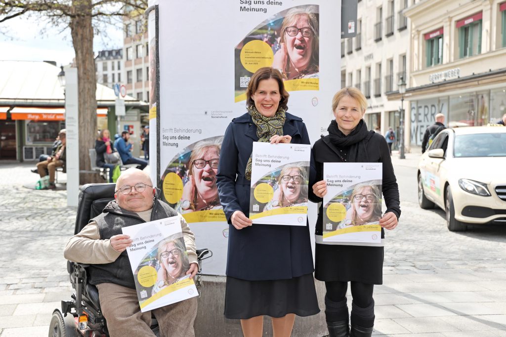 Ein Mann im Rollstuhl und zwei Frauen stehen vor einer Litfaß-Säule auf dem Münchner Viktualienmarkt. Die drei Personen sind Oswald Utz, ehrenamtlicher Behindertenbeauftragter der Landeshauptstadt München, Bürgermeisterin Verena Dietl und Sozialreferentin Dorothee Schiwy.. Alle drei halten ein Plakat in der Hand. Auf dem Plakat steht ""Betrifft Behinderung - Sag uns deine Meinung". Dieses Plakat hängt auch auf der Litfaß-Säule im Hintergrund.