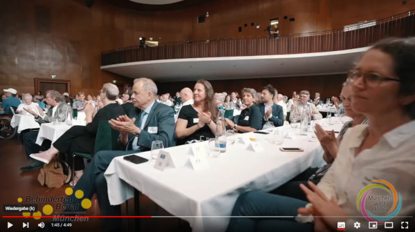 Menschen sitzen an langen Tischen in einem Saal und klatschen.