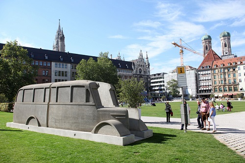 Bus-Silhouette aus grauem Beton steht auf dem Marienhof, daneben einige Menschen.