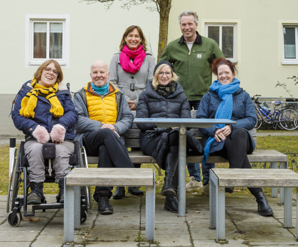 Sechs Menschen haben sich um einen Tisch und zwei Sitzbänke gruppiert. Sie sind mittleren Alters. Von den vier Frauen steht eine, zwei sitzen auf einer Bank und eine Frau sitzt im Rollstuhl. Von den zwei Männern sitzt einer auf der Bank, der andere steht.