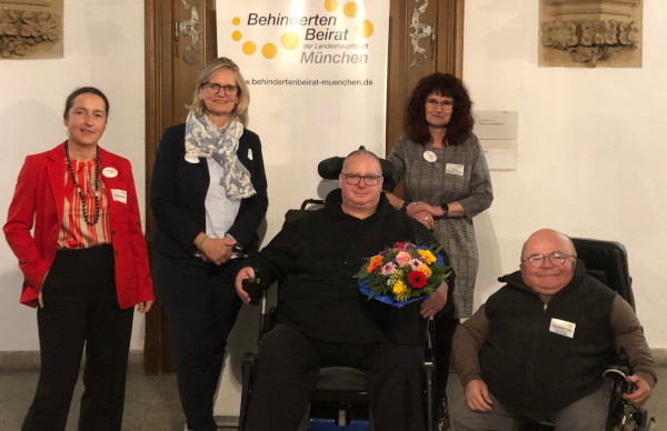 Drei weibliche Personen, zwei männliche Personen im Rollstuhl. Der Mann in der Mitte hält einen Blumenstrauß in der Hand. Im Hintergrund ein Banner mit der Aufschrift Behindertenbeirat der Landeshauptstadt München